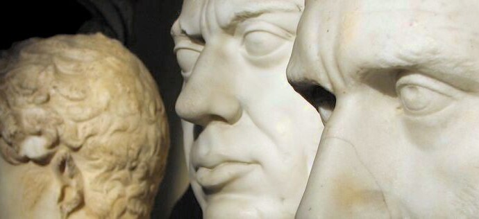 Two marble faces of men in the foreground. There is another statue in the background.