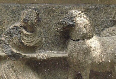 A stone relief. Three people are pulling a horse on wheels. A woman looks at the scene.