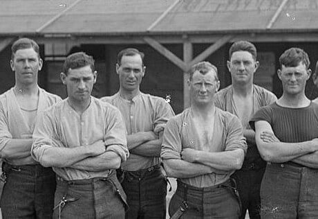 Eight young, strong soldiers wearing casual clothes stand in a yard with their arms crossed.