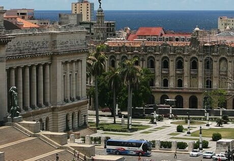 A large, monumental city square. It is surrounded by European-style buildings. The ocean is in the background.