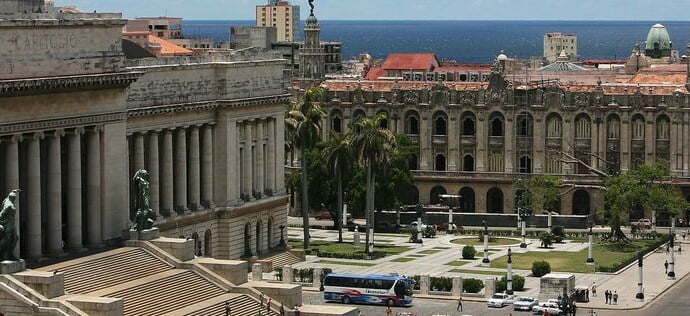A large, monumental city square. It is surrounded by European-style buildings. The ocean is in the background.
