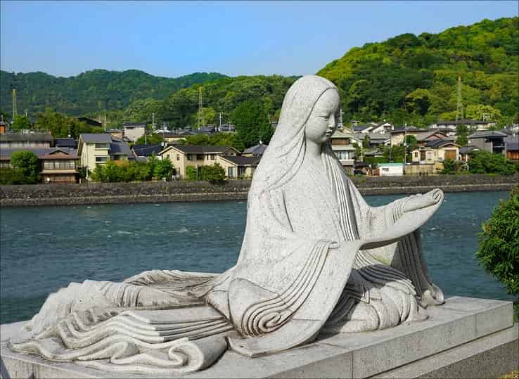 Outdoors, next to a river. Gray stone statue of a woman. She sits reading a scroll.