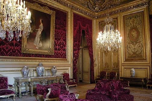 Luxurious room decorated in gold and burgundy. With paintings, teardrop chandeliers, and velvet furniture.