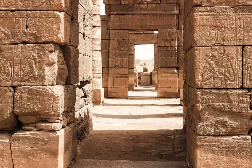 A sequence of archways leading to a stone temple. The columns are carved with scenes.