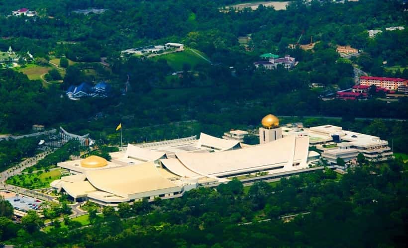 Aerial view of a massive building with white curved roofs and golden domes. It is surrounded by trees.