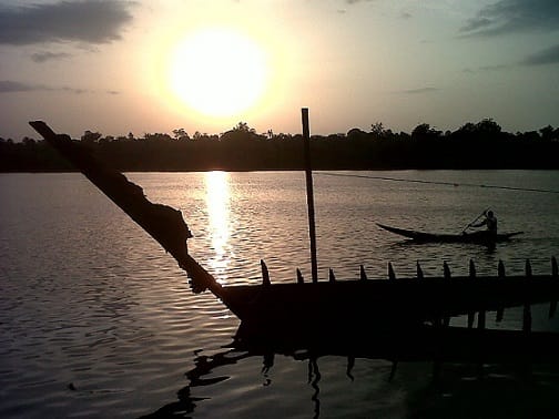 Sunset over a estuary. A canoe crosses the water. On the background, the shore has dense vegetation.