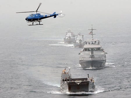 Four warships in formation with a helicopter hovering above. Open sea.