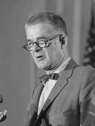 Black and white photo of a man talking at a press conference. He is dressed in a suit and wears glasses.