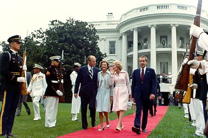 Two couples walk away from the White House that is seen in the background. They are flanked by military personal in parade dress.