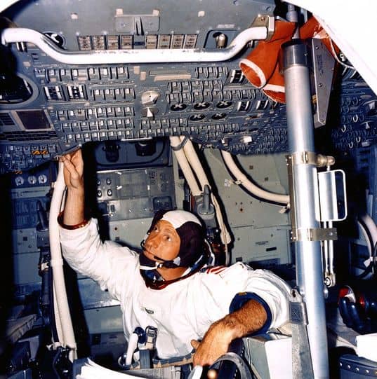A man wearing a partial astronaut's suit sits inside a small cockpit full of buttons.