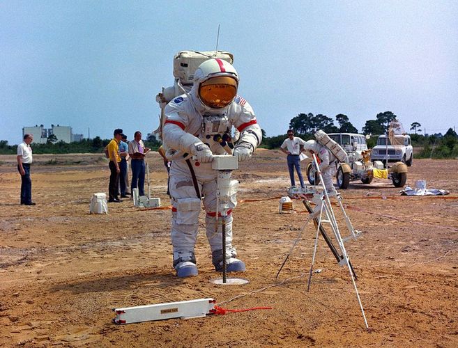 An astronaut wearing his suit on Earth, on a field. He is drilling the ground.