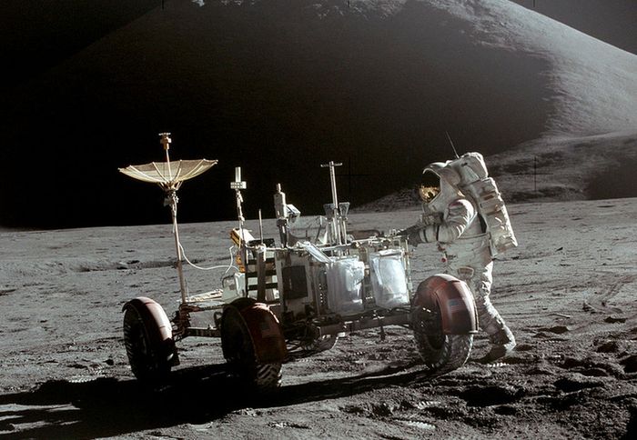 Outside. Picture on the Moon's surface. A suited astronaut approaches the rover. The ground is gray and the sky is black.