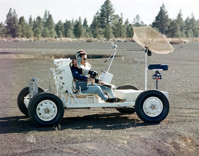 Outdoors on Earth. Two men sit on the rover during a training exercise. The rover is a low, small, open, simple car with four small wheels and seats for two.