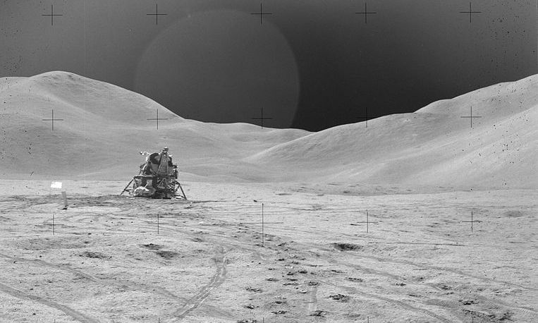 Lunar surface. The lunar module sits on the ground. There are mountains on the background.