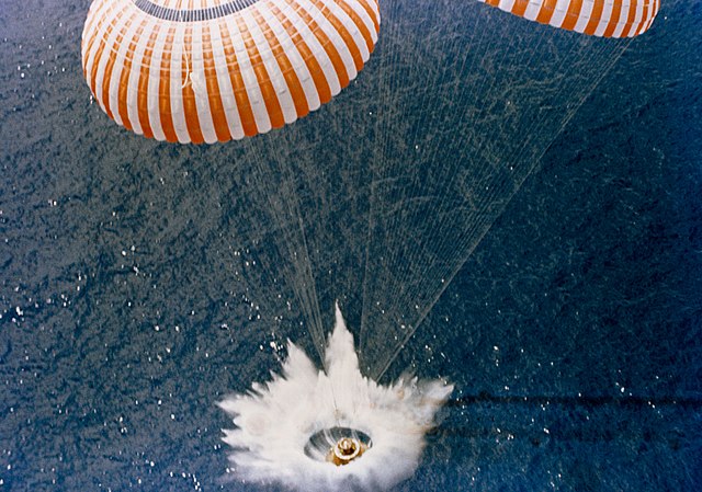An object splashes down in the middle of the ocean. It is attached to two open parachutes.
