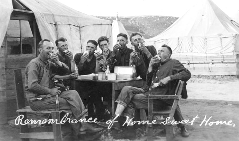 Six young soldiers sit outside around a table and have some fun. They are smiling and eating.