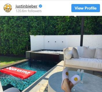 Justin has taken a picture of his backyard. It has a pool, grass and some modern furniture. His feet and slippers are seen in the shot.