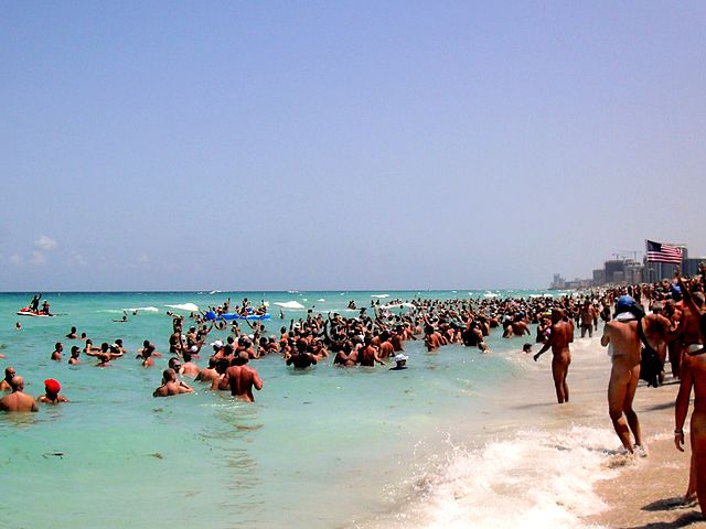 A sunny beach. Dozens of nudists are bathing in the sea.