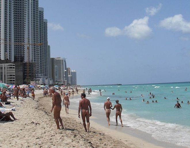 A sunny beach with green waters. Nudists walk along the beach, there are tall buildings in the background.