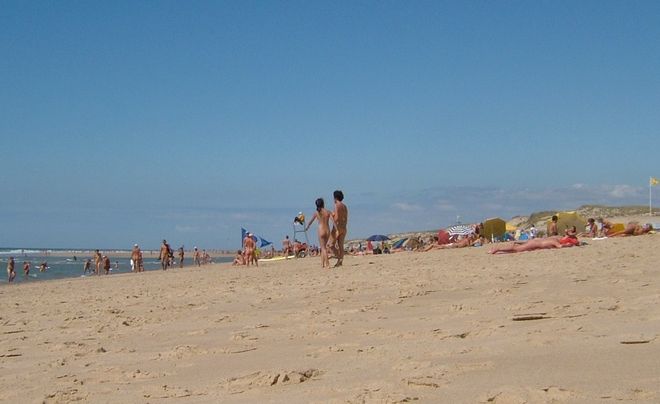A wide, sunny beach with lots of room. There are many nudists walking and sunbathing.