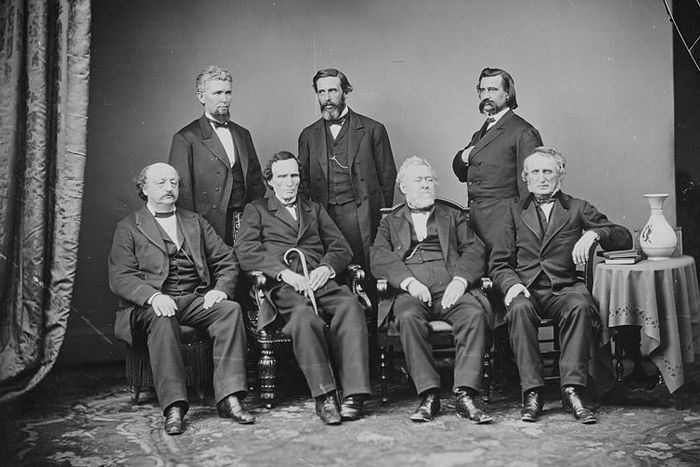 Black and white photograph of 7 seated men elegantly dressed.