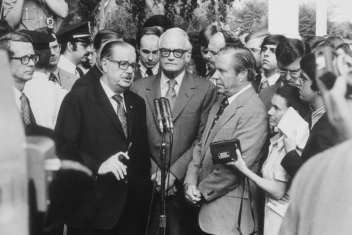 Black and white photo. Three men talk during a press conference outdoors. They are surrounded by journalists.