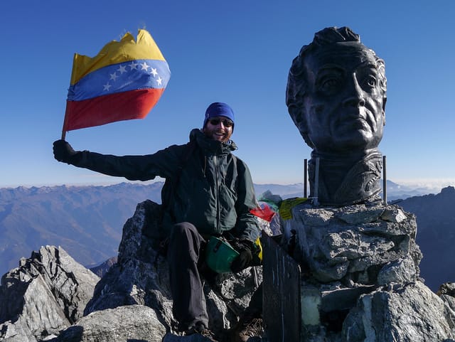 A man stands on top of a mountain and waves a Venezuelan flag. Next to him, on the peak, is a statue of Bolivar.