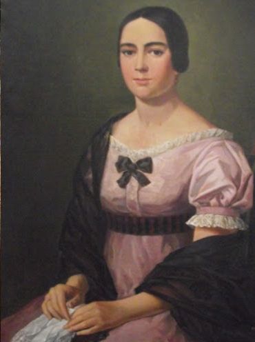 A woman wearing a pink dress and a black shawl. She had dark hair, her face is round, and she has big eyes
