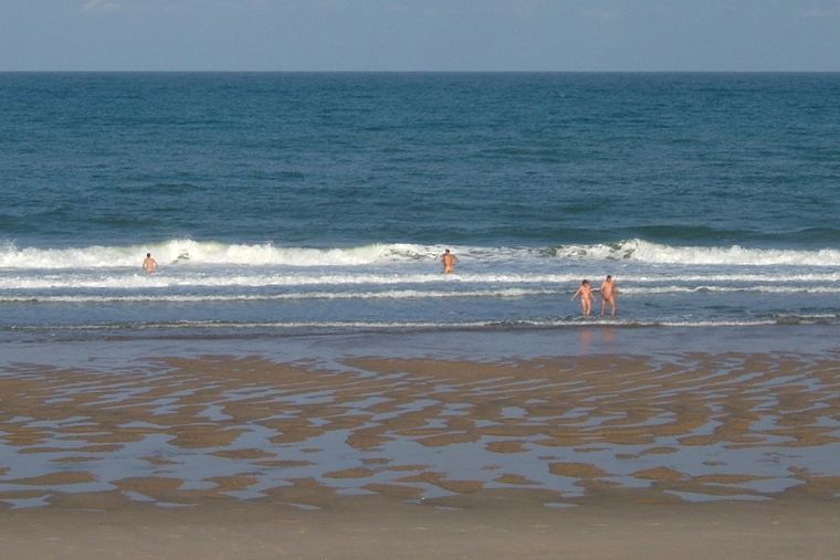 A wide beach. Some nudists are swimming in the ocean.