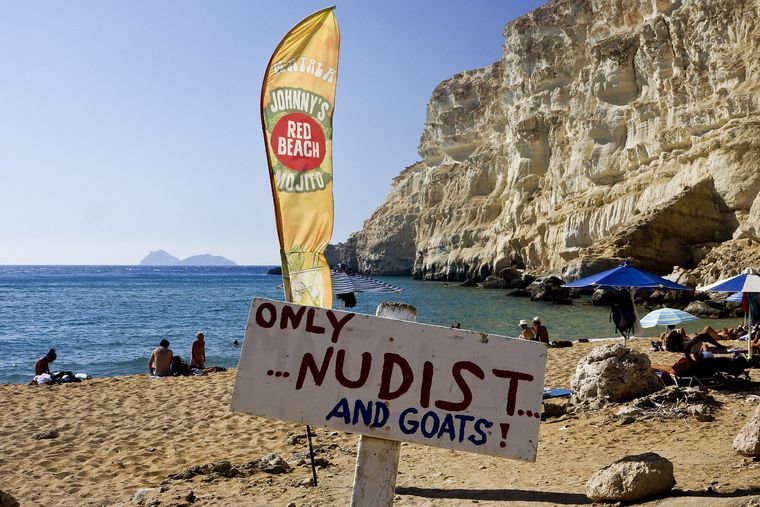 A beach surrounded by cliffs. There is a sing that reads: "Only nudists... and goats"