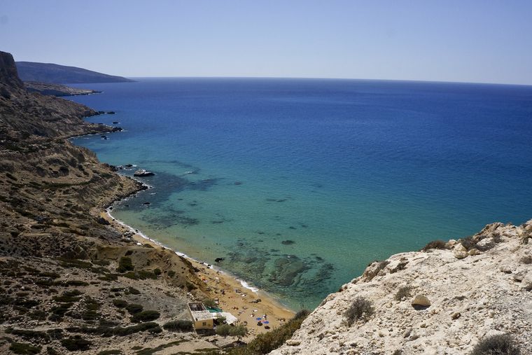 The nudist beach seen from above, from the cliff. The water is blue and the sand is red.