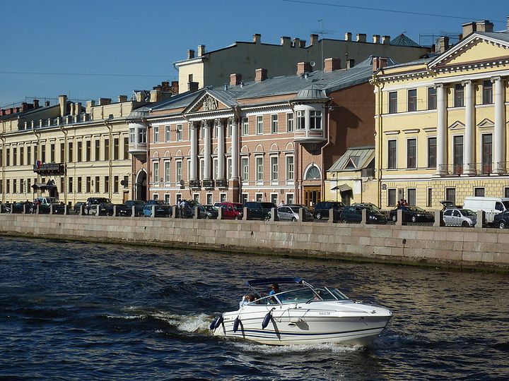 A yacht navigating a canal. The canal's bank has many neoclassical buildings.