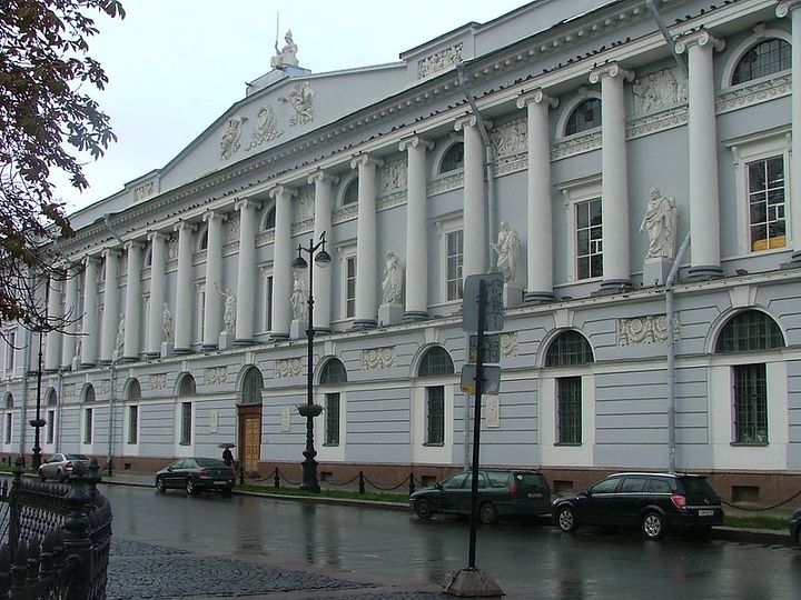 A greyish Neoclassical building seen from the street. It has many decorative columns along its facade.