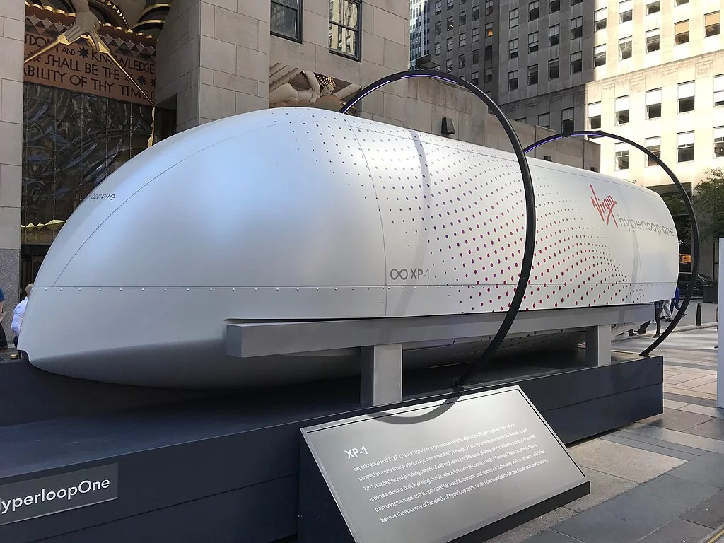 Picture of a small pod exhibited on a city square. It looks like the front tip of a train and probably seats some 20 people.