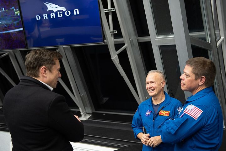 Picture of Elon, in a suit, talking to two astronauts that are wearing their uniform.