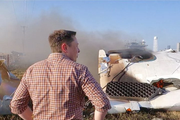 Picture outdoors. Elon looking at a big crashed spacecraft.