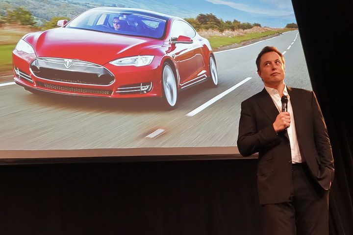 Picture of Elon Musk standing on stage in a suit and holding a microphone. Behind him is a big screen which shoes a red car.