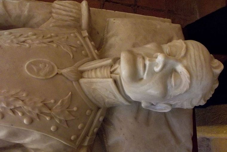 Face of a white marble statue. The statue is lying down; looks like a sleeping man. His face is thin, long, and bony. He has prominent high cheekbones, a prominent long, thin nose, and a prominent chin. His eyebrows are bushy and arched, high in his forehead. His closed eyes are big and have big eyelids. His lips are thin, and his forehead is large.