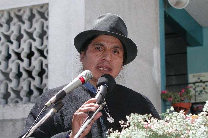 A man in traditional clothes and hat gives a speech outdoors.