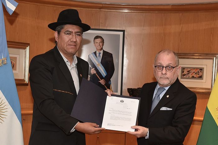 Two men hold a diploma. Diego is tall and wears traditional clothes including a hat. The other is shorter and wears a suit.