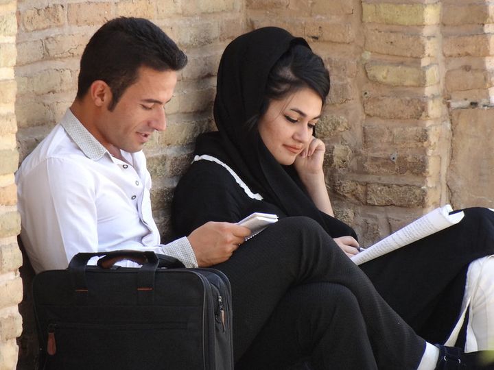 A couple, a handsome man and a woman dressed in black, sit against a stone monument, reading and smiling.