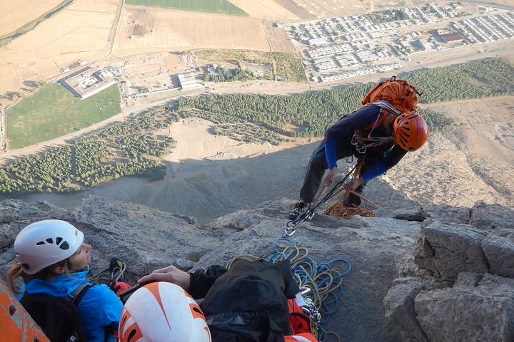 Four rock climbers. Three wait on top of a mountain while another makes his way up.