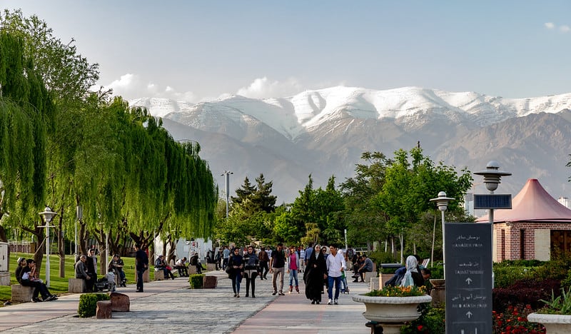 A park. There are tall trees on the left, mountains in the background, and in the foreground people walk on a lane.