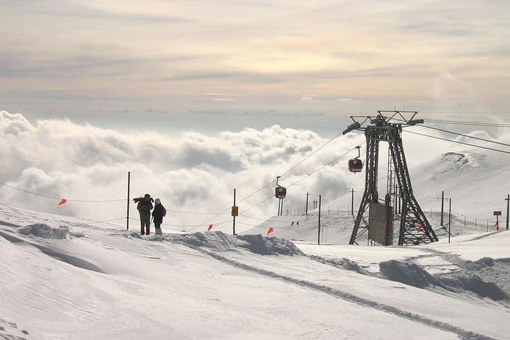 On top of big mountains covered with snow. Two persons stand on the top of the mountain. There are clouds below them. There is a gondola lift.