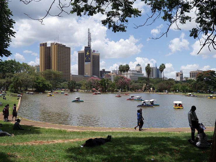 A park with a lake. A couple strolls along the lake, and Nairobi's skyline can be seen in the back.