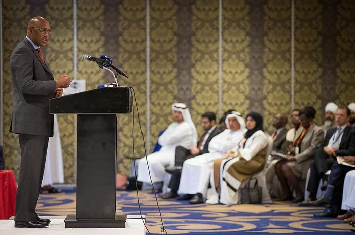 Inside an auditorium. On the left is a suited man who stands behind a podium giving a speech. On the right are the representatives of the UAE sitting down, listening.