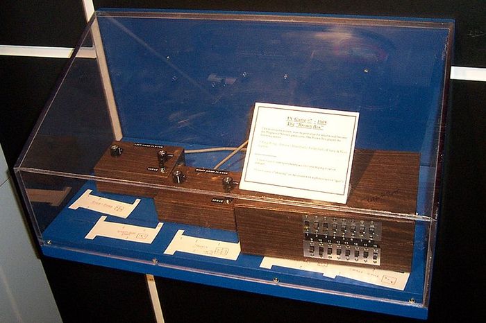 A museum display. Inside a glass box are a brown box with many switches and two brown controllers.