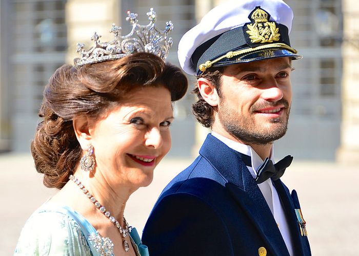 Picture of a smiling woman who is elegantly dressed and wears a crown. She is walking next to a younger man who is wearing his formal military uniform.