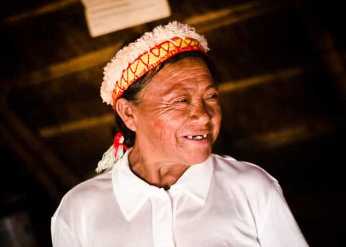 An older woman smiling, wearing traditional clothes inside a hut