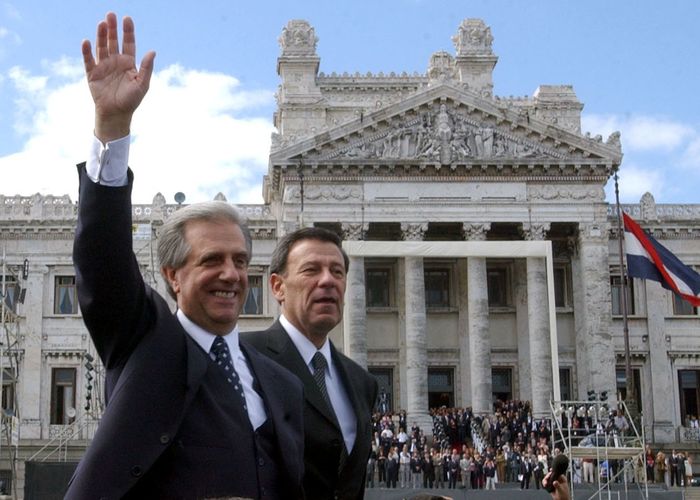 Two man in suits walking through a square with beautiful buildings. They are waving to the crowd.
